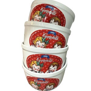 May include: A stack of four white ceramic Campbell's soup bowls. Each bowl features a red oval design with the Campbell's logo, the year 2000, and illustrations of children. The bowls are stacked on top of each other.