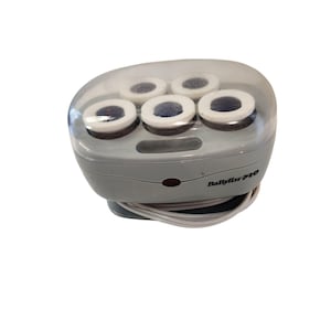 May include: A grey BaBylissPRO hair setter unit with a translucent lid, containing five cylindrical hair rollers with white rims. The device features a small red indicator light on the front and includes a white power cord coiled at the base.