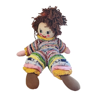 May include: A handmade rag doll with brown yarn hair and a painted face. The doll wears a patchwork outfit with pink, blue, yellow, and green fabric. The doll's arms and legs are pink and brown.