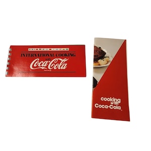 May include: A red spiral-bound cookbook titled International Cooking with Coca-Cola featuring a row of international flags and a red vertical pamphlet titled Cooking with Coca-Cola showing a plate of food with potatoes and meat.