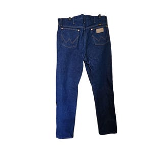 K&ouml;nnte beinhalten: Dunkelblaue Jeans mit dem Wrangler-Logo auf der Ges&auml;&szlig;tasche. Die Jeans h&auml;ngen an einem schwarzen Kleiderb&uuml;gel. Die Jeans haben ein klassisches F&uuml;nf-Taschen-Design und ein gerades Bein.