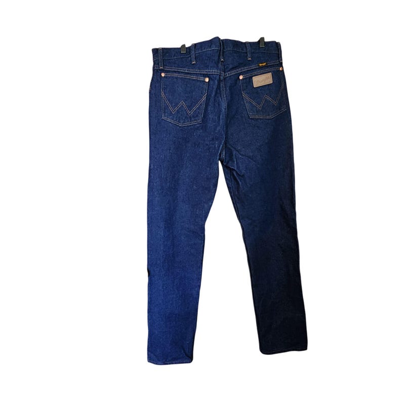 K&ouml;nnte beinhalten: Dunkelblaue Jeans mit dem Wrangler-Logo auf der Ges&auml;&szlig;tasche. Die Jeans h&auml;ngen an einem schwarzen Kleiderb&uuml;gel. Die Jeans haben ein klassisches F&uuml;nf-Taschen-Design und ein gerades Bein.