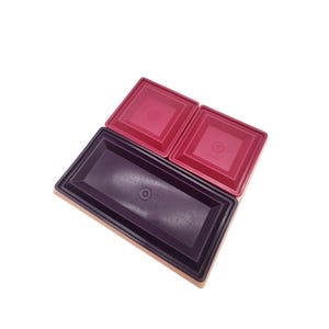 May include: An overhead shot of three plastic containers. Two are square with a deep red hue, and one is rectangular with a dark purple color. All have a circular indentation in the center of the lid.