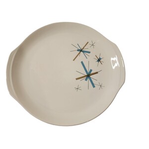 May include: A round, off-white ceramic serving platter with integrated side handles. The surface features a graphic pattern of intersecting lines in black, light blue, and metallic gold, arranged in a starburst motif on the right side.