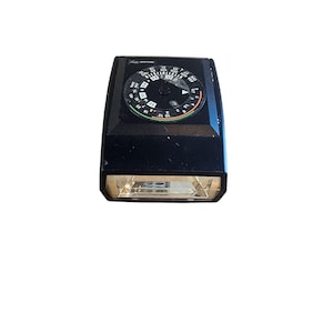 May include: A black vintage light meter with a circular dial featuring numbers and markings. The meter has a rectangular shape with a clear lens at the bottom. The brand name "Sekonic" is visible on the top.