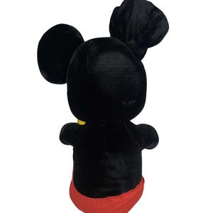 May include: A plush Mickey Mouse toy, viewed from the back, featuring a black body, ears, and head, with red shorts and a yellow collar. The toy is made of soft, velvety material.