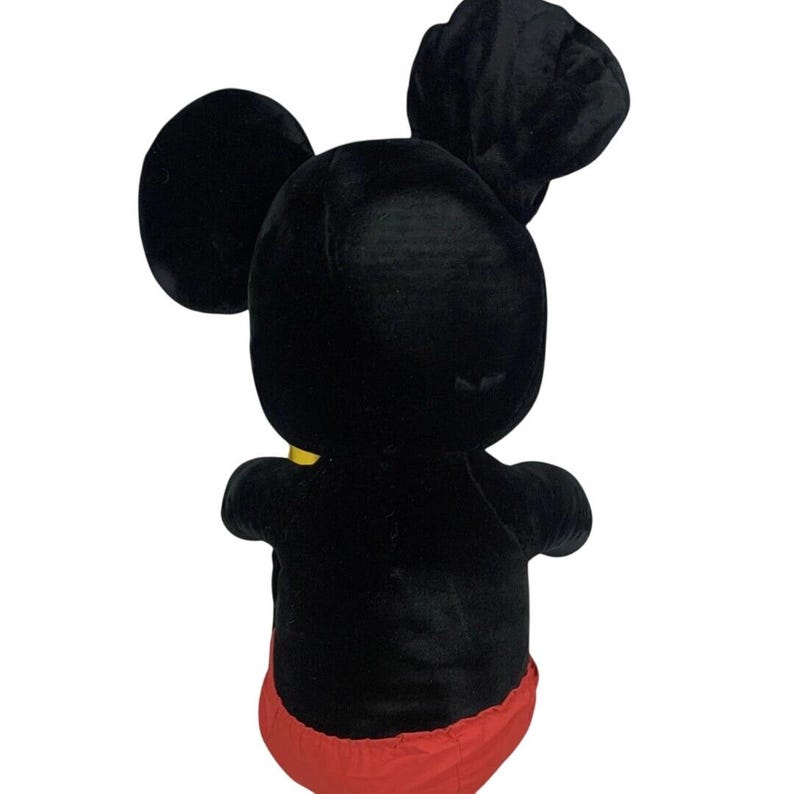 May include: A plush Mickey Mouse toy, viewed from the back, featuring a black body, ears, and head, with red shorts and a yellow collar. The toy is made of soft, velvety material.