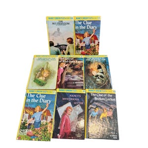 May include: A collection of Nancy Drew mystery books with yellow spines and colorful covers. Titles include "The Sky Phantom," "The Clue in the Diary," and "The Secret of Red Gate Farm." The books are arranged on a white surface.