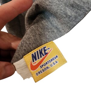 May include: Close-up of a gray t-shirt with a yellow Nike tag. The tag features the Nike logo in orange and the words "SPORTWEAR OREGON, U.S.A."