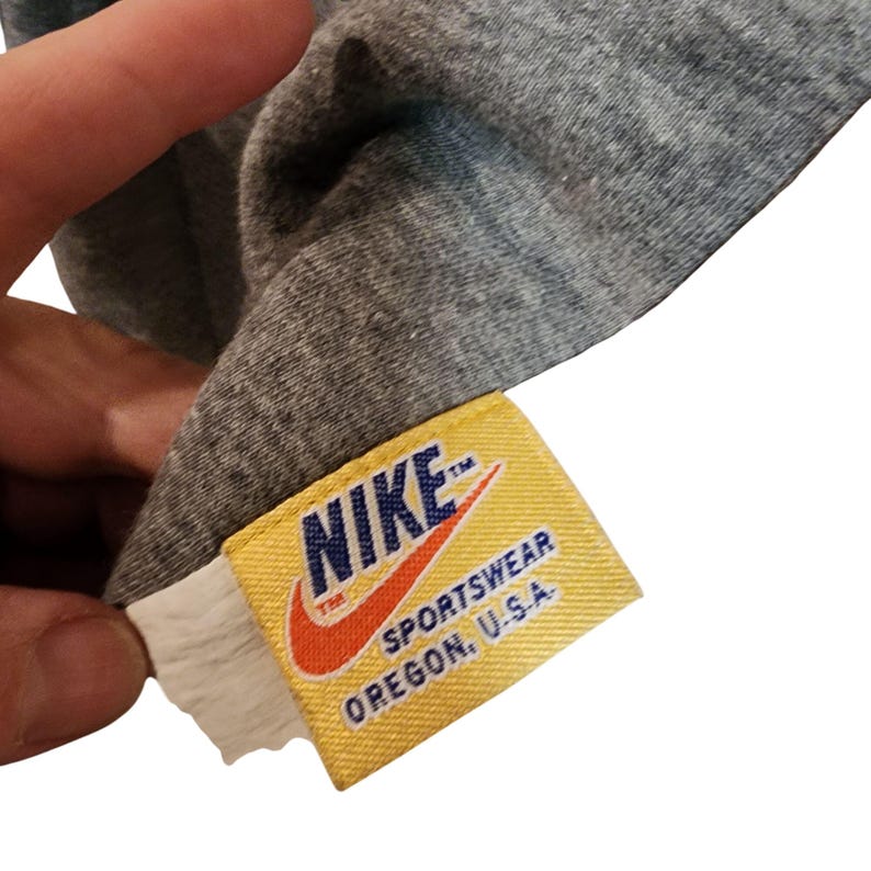 May include: Close-up of a gray t-shirt with a yellow Nike tag. The tag features the Nike logo in orange and the words "SPORTWEAR OREGON, U.S.A."