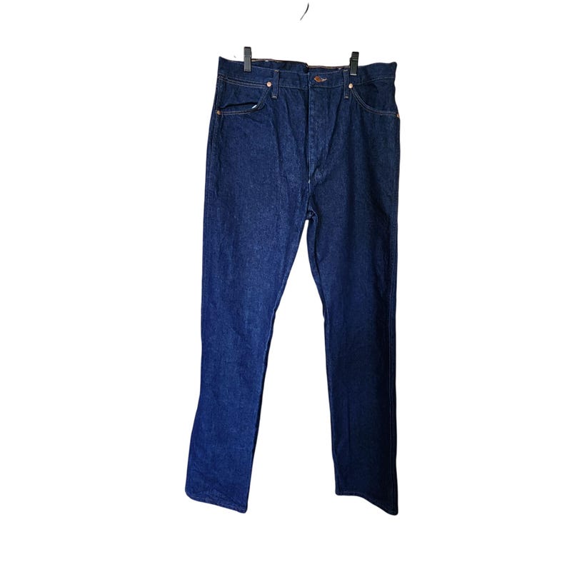 K&ouml;nnte beinhalten: Dunkelblaue Jeans aus Denim, die an einem schwarzen Kleiderb&uuml;gel h&auml;ngen. Die Jeans haben ein klassisches F&uuml;nf-Taschen-Design mit kupferfarbenen Nieten und einem Knopfverschluss. Der Stoff scheint ein mittelschwerer Denim zu sein.