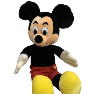 May include: A large plush Mickey Mouse toy. The iconic character has a black body, red shorts, yellow shoes, and white gloves. Mickey's face is tan with a black nose and large black ears. The toy is designed to sit upright.