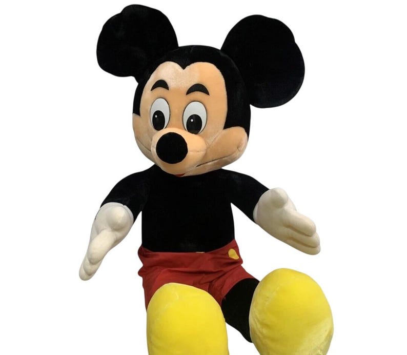 May include: A large plush Mickey Mouse toy. The iconic character has a black body, red shorts, yellow shoes, and white gloves. Mickey's face is tan with a black nose and large black ears. The toy is designed to sit upright.