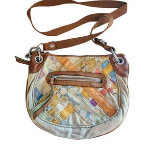 May include: A cream-colored crossbody bag with a patchwork design featuring floral patterns and geometric shapes in various colors. The bag has brown leather trim, a zippered front pocket, and a brown adjustable strap. The bag measures approximately 10 inches wide.