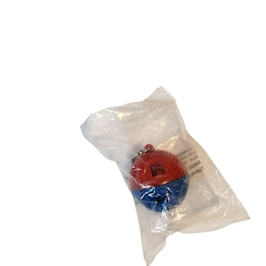 May include: A red and blue bell-shaped keychain inside a clear plastic bag. The keychain has a small silver chain and a square opening on the red top. The plastic bag has some text printed on it.