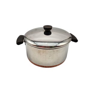 May include: Stainless steel cooking pot with a lid and two side handles. The lid has a dark brown knob. The pot sits on a round, light brown base. The pot is round and has a shiny, metallic finish.