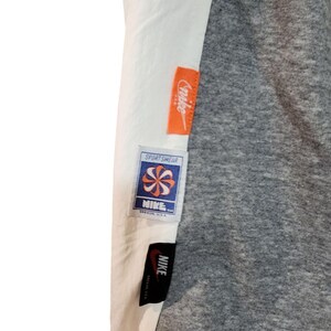 May include: Close-up of a gray and white garment with multiple Nike tags. The tags are orange, white, and blue, with the Nike logo and the word "Sportswear" visible. The garment has a gray, textured appearance.
