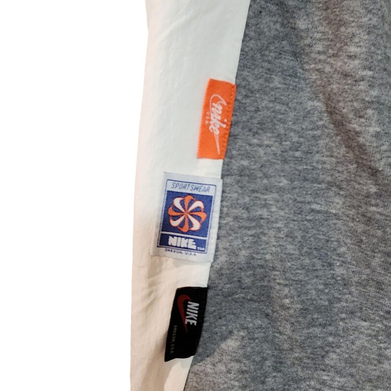 May include: Close-up of a gray and white garment with multiple Nike tags. The tags are orange, white, and blue, with the Nike logo and the word "Sportswear" visible. The garment has a gray, textured appearance.