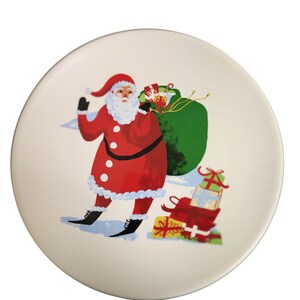 May include: A white ceramic plate with a Christmas design featuring Santa Claus carrying a green bag full of presents. Santa is wearing a red suit with white trim and a white beard. The plate is decorated with red and green accents.
