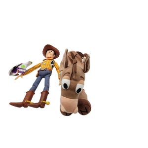 May include: A plush Woody doll in a cowboy hat, yellow plaid shirt, blue jeans, and brown boots, holding a Buzz Lightyear toy. Beside it is a plush brown horse head with large, expressive eyes. Toy Story themed.
