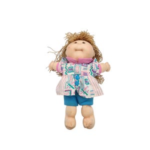 May include: A vintage doll with a soft, round face and light brown hair. The doll wears a patterned top with blue buttons, a pink collar, and blue shorts. The doll's arms and legs are made of soft material.