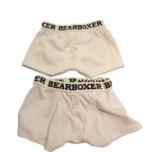 May include: Two pairs of white boxer shorts with black text that reads "BEARBOXER" on the waistband. One pair has a button closure. The shorts are made of a soft, light-colored fabric.