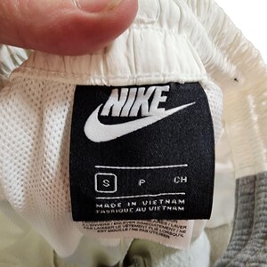 May include: Close-up of a black Nike tag with a white Nike logo and text. The tag reads "MADE IN VIETNAM" and "FABRIQUE AU VIETNAM". The tag is attached to a pair of light-colored athletic shorts.