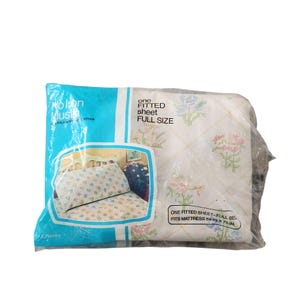 May include: A packaged, full-size fitted sheet. The sheet is white with a floral pattern in pink, blue, and green. The packaging is a clear plastic bag with blue and white text that reads "No Iron Muslin" and "One Fitted Sheet Full Size". The sheet fits a 54 in x 75 in mattress.