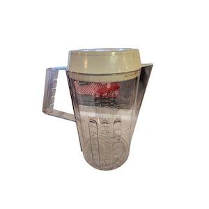 May include: A clear plastic blender pitcher with a white lid and a handle. The pitcher has measurement markings on the side.
