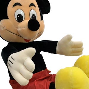 May include: A large plush Mickey Mouse toy. The iconic character is depicted with a black body, red shorts, yellow shoes, and white gloves. The toy has a smiling expression and outstretched arms, inviting a hug.