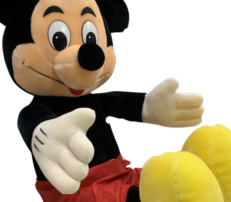 May include: A large plush Mickey Mouse toy. The iconic character is depicted with a black body, red shorts, yellow shoes, and white gloves. The toy has a smiling expression and outstretched arms, inviting a hug.