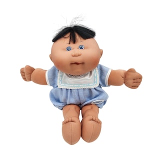 May include: A Cabbage Patch Kids doll with a soft, vinyl face, blue eyes, and black hair. The doll is wearing a light blue knit outfit with a white bib and is posed with arms outstretched.