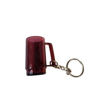 May include: A burgundy-colored miniature mug keychain. The small mug has a handle and is attached to a gold-colored chain and keyring. The keychain is made of a translucent material.