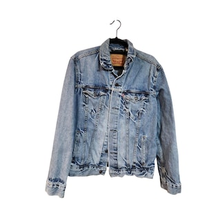 May include: Light blue denim jacket with a classic collar, button-up front, and two chest pockets. The jacket has a distressed look with faded areas and a red tag on the chest pocket. It is hanging on a black hanger.