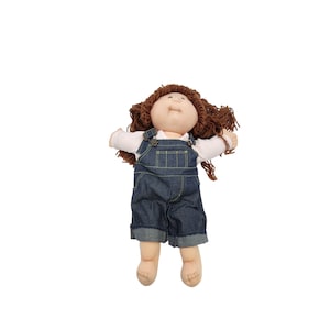 May include: A vintage doll with a soft, fabric body, wearing a white long-sleeved shirt and denim overalls. The doll has brown yarn hair and a smiling face. The overalls have metal clasps and rolled-up cuffs.