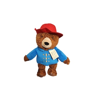 May include: A plush brown bear wearing a blue jacket and a red hat. The bear has a tag that says "Paddington Bear, Love from Peru".