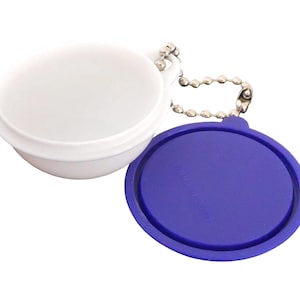 May include: A white, round container with a blue lid, attached to a silver ball chain. The container is open, revealing its interior. The lid is a deep blue color and has a circular shape.