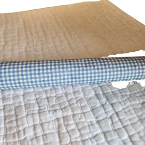 May include: A roll of blue and white gingham patterned paper or fabric placed between two layers of white quilted material. The quilted fabric features a grid-like stitched texture, while the central roll displays a classic small check pattern.