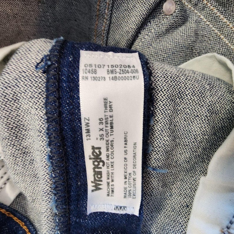 K&ouml;nnte beinhalten: Nahaufnahme eines Wrangler Jeansetiketts. Das wei&szlig;e Etikett zeigt schwarzen Text, einschlie&szlig;lich der Marke, der Gr&ouml;&szlig;e 35 x 36 und Pflegehinweise. Die Jeans ist aus dunkelblauem Denim mit sichtbaren N&auml;hten und einem Knopf.