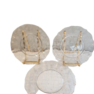 May include: Three clear glass plates featuring a delicate etched floral pattern along the scalloped edges. Each plate has a smooth circular center and a textured rim. Two of the plates are supported by gold-toned metal display stands.