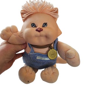 May include: A plush pink and white cat wearing blue overalls and a brown collar with a gold tag that says "Bazzook".