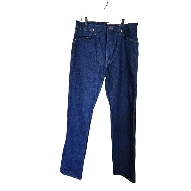 K&ouml;nnte beinhalten: Dunkelblaue Jeans aus Denim, die an einem Kleiderb&uuml;gel h&auml;ngen. Die Jeans haben ein klassisches F&uuml;nf-Taschen-Design mit kupferfarbenen Nieten und einem Knopfverschluss. Der Stoff scheint ein mittelschwerer Denim zu sein.