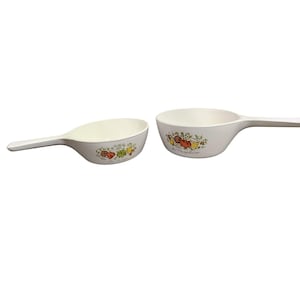 May include: Two white ceramic kitchen bowls with handles. The bowls have a floral design with red, yellow, and green fruits and the words "Le Marché" and "La Cuisine" printed on them.