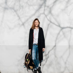 May include: A woman wearing a black coat, white shirt, and blue jeans stands in front of a white wall with a tree shadow. She is holding a black backpack.