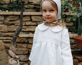 May include: A long-sleeved white dress featuring a rounded Peter Pan collar and a gathered waistline, paired with a matching light gray fabric bonnet that ties under the chin with thin ribbons, creating a coordinated and simple ensemble.