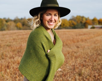 May include: A woman wearing a dark green hat and a green shawl, smiling in a field. The shawl is draped over a beige dress. The background features a field of dry grass and a cloudy sky.