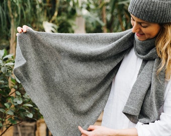 May include: A grey knitted scarf with a triangular shape. The scarf is being held by a person wearing a white shirt and a grey beanie.