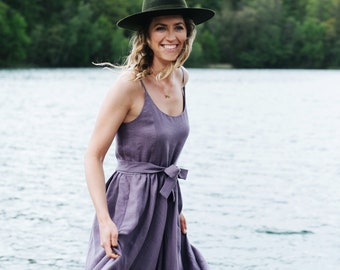 May include: A woman wearing a purple linen dress with a wide belt and a green fedora hat. She is standing on a wooden dock by a lake.