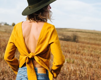 Peut inclure: Une femme portant un chemisier jaune moutarde noué dans le dos, un jean bleu et un chapeau fedora vert foncé. Elle se tient dans un champ d'herbe sèche.