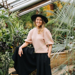 May include: A woman wearing a pink sweater and black wide-leg pants stands in a greenhouse. She is wearing a wide-brimmed hat and smiling at the camera.